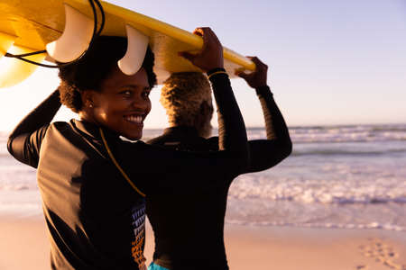 Portrait of smiling african american mature woman with man carrying surfboard on head at beach. sunset, nature, unaltered, love, togetherness, enjoyment, aquatic sport, holiday, active lifestyle.の写真素材