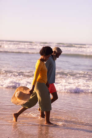 Side view of happy african american couple walking at shore against clear sky at sunset, copy space. nature, hat, unaltered, love, togetherness, retirement, enjoyment and holiday concept.の写真素材