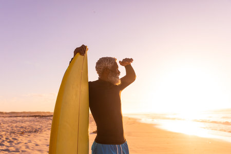 African american bearded senior man with surfboard shielding eyes while looking at sea at sunset....