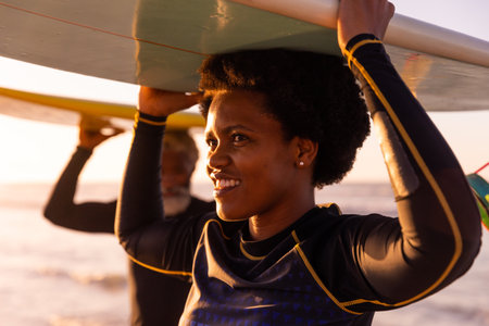 African american mature woman and senior man carrying surfboards on heads at beach at sunset....
