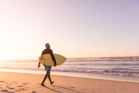 African american senior man with surfboard walking at beach against clear sky at sunset, copy space. sunlight, nature, unaltered, retirement, aquatic sport, holiday and active lifestyle concept.の写真素材
