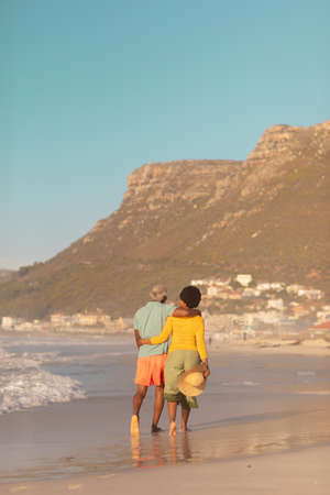 Rear view of african american couple with arms around walking at beach against mountain and blue sky. copy space, nature, hat, unaltered, love, togetherness, retirement, enjoyment and holiday.の写真素材