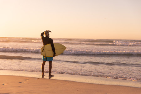 Rear view of african american senior man carrying surfboard looking at sea against clear sky. sunset, copy space, nature, unaltered, retirement, aquatic sport, holiday and active lifestyle concept.の写真素材