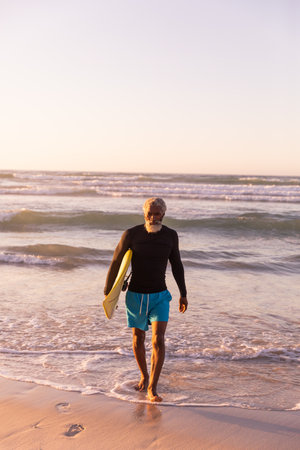 Full length of bearded african american senior man with surfboard walking against sea and clear sky. copy space, sunset, nature, unaltered, retirement, aquatic sport, holiday and active lifestyle.の写真素材