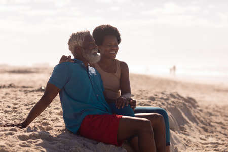 African american bearded senior man and mature woman with afro hair sitting on beach against sky. happy, summer, copy space, nature, unaltered, love, togetherness, retirement, enjoyment and holiday.の写真素材