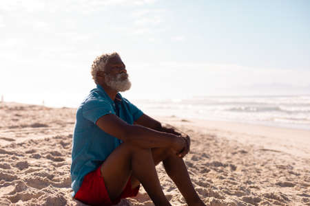 Thoughtful bearded african american senior man with gray hair sitting on sandy beach against sky....