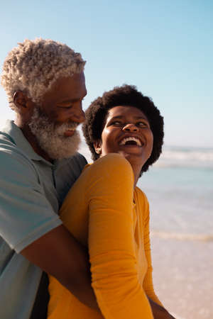 Bearded african american senior man embracing mature woman with afro hair at beach against clear sky. copy space, romance, cheerful, unaltered, love, togetherness, retirement, enjoyment and holiday.の写真素材