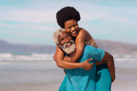Playful bearded african american senior man piggybacking happy mature woman against sea and sky. summer, nature, happy, afro hair, unaltered, love, togetherness, retirement, enjoyment and holiday.の写真素材