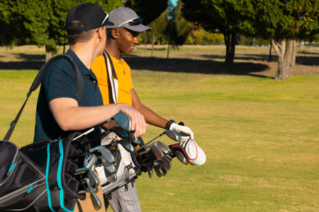 Side view of multiracial young male friends wearing caps with golf bags walking at golf course....