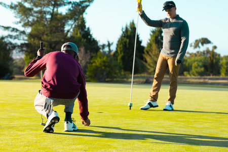 Caucasian young man holding flag while playing golf with african american friend at golf course. summer, golf, unaltered, friendship, togetherness, nature, sport and weekend activities concept.の写真素材