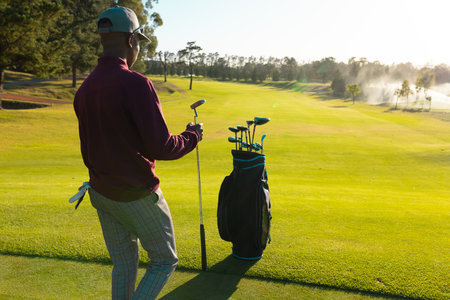African american young man with golf clubs in bag walking on golf course against sky in summer. summer, copy space, green, golf, unaltered, nature, sport and weekend activities concept.の写真素材