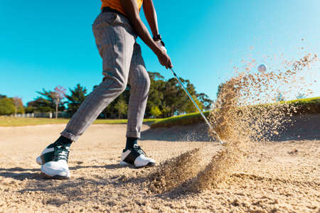 Low section of african american young man hitting golf ball with club on sand against clear sky. copy space, golf course, golf, unaltered, nature, sport and weekend activities concept.の写真素材