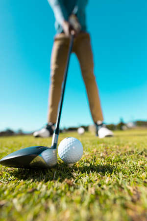 Low section of african american young man aiming golf ball with club against clear sky in summer. copy space, low angle view, golf, golf course, unaltered, sport and weekend activities concept.の写真素材