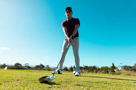 Low angle view of caucasian young man aiming ball with golf club while standing against clear sky. copy space, blue, summer, golf, unaltered, nature, sport and weekend activities concept.の写真素材