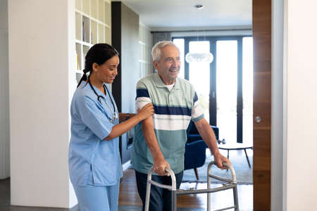 Biracial female physiotherapist assisting happy caucasian senior man in walking with walker at home....