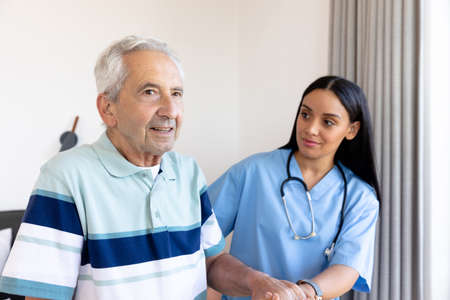 Biracial female physiotherapist holding hand and assisting caucasian senior man in standing at home. Copy space, unaltered, physical therapy, support, healthcare, patient, retirement and recovery.の写真素材