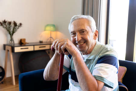 Portrait of smiling caucasian senior man with walking cane sitting on chair in living room at home. Unaltered, physical impairment, retirement lifestyle and disability concept.の写真素材
