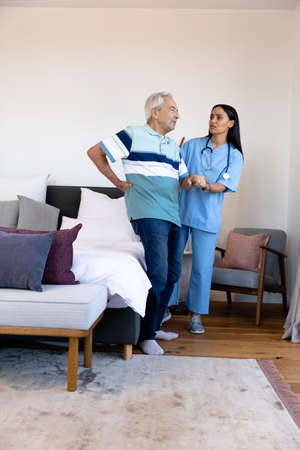 Biracial female physiotherapist assisting caucasian senior man with backache in standing at home....