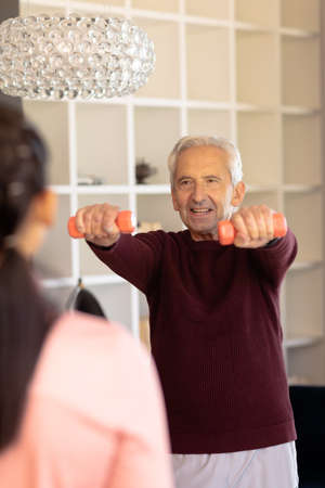 Caucasian senior man exercising with dumbbells while looking at biracial female physiotherapist. Home, unaltered, physical therapy, exercise, healthcare, retirement, treatment and recovery concept.の写真素材