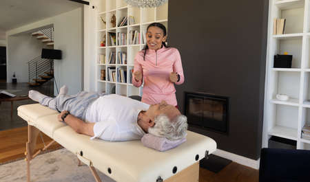 Biracial female physiotherapist gesturing while guiding caucasian senior man lying on table at home....