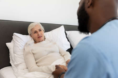 African american male health worker supporting caucasian senior woman lying on the bed at home....
