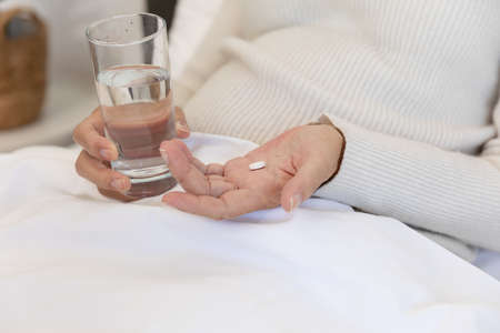 Mid section of caucasian senior woman holding a glass of water and medical pill lying on bed. Medical care and retirement senior lifestyle conceptの写真素材