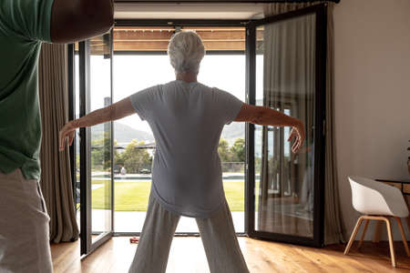 Rear view of caucasian senior woman performing stretching exercise at home. Physiotherapy and...