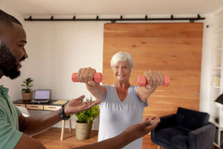 African american male physiotherapist helping caucasian senior woman to do exercise with dumbbells. Physiotherapy and rehabilitation conceptの写真素材