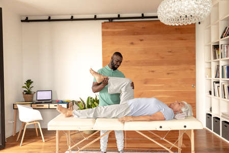 African american male physiotherapist giving leg massage therapy to caucasian senior woman at home....