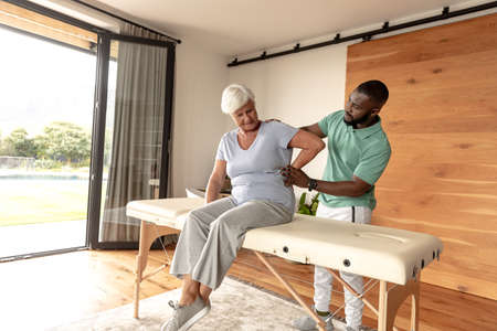African american male physiotherapist giving back massage therapy to caucasian senior woman at home....