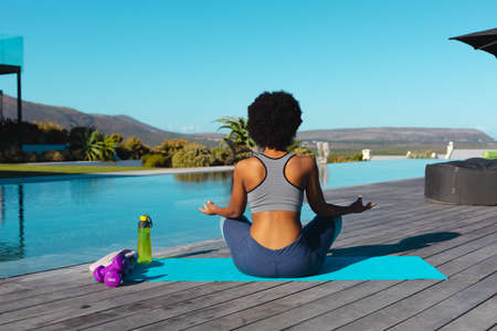 Rear view of african american woman practicing yoga and meditating near the pool. Fitness and...
