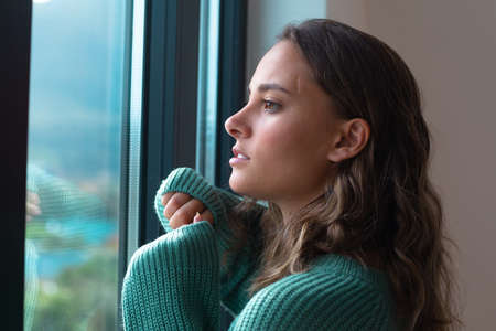 Caucasian beautiful young woman looking out of the window at home. Self-care and relaxation concept