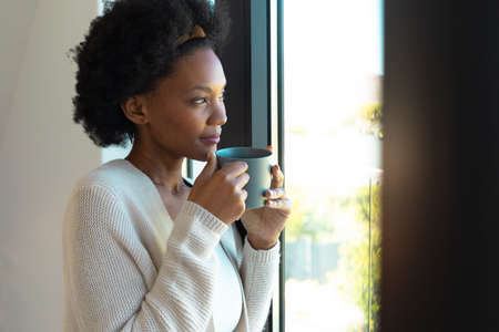 African american young woman holding a coffee cup looking out of the window at home. Relaxation...