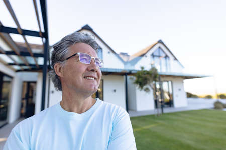 Smiling caucasian senior man wearing eyeglasses looking away against house in yard, copy space....
