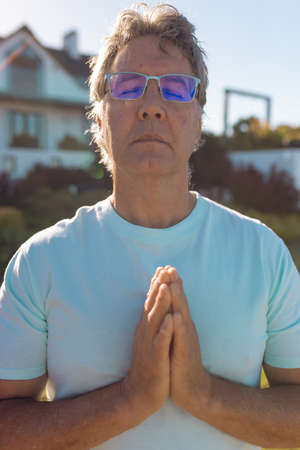 Caucasian senior man wearing eyeglasses with eyes closed meditating in prayer position in yard....