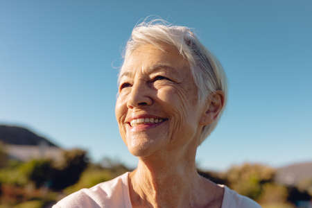 Close-up of cheerful asian senior woman with short hair looking away against clear blue sky in yard....