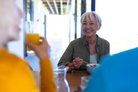 Happy asian senior woman looking at multiracial friends talking while having breakfast at table....
