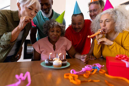 Multiracial friends singing and blowing party horns while senior woman blowing birthday candles. Nursing home, cupcake, enjoyment, unaltered, togetherness, support, assisted living, retirement.の写真素材