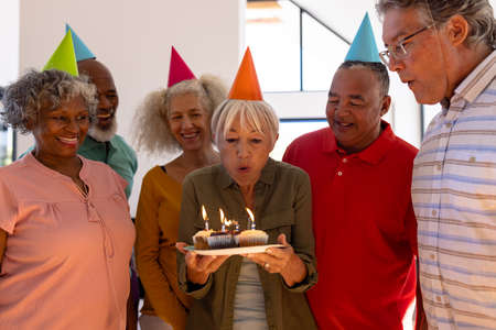 Happy multiracial friends wearing party hats looking at senior woman blowing birthday candles. Cupcake, nursing home, enjoyment, unaltered, togetherness, support, assisted living and retirement.の写真素材