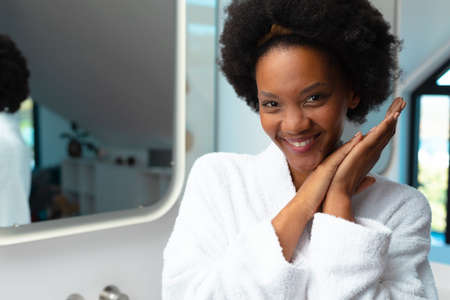 Portrait of smiling african american young afro woman wearing bathrobe in bathroom, copy space. Unaltered, spa, home, relaxation, pampering, detox, body care and wellbeing concept.の写真素材