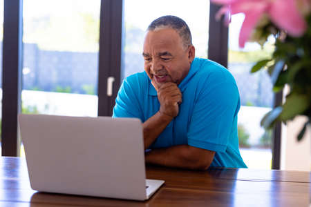 Smiling biracial senior man with hand on chin watching video over laptop while sitting at table....