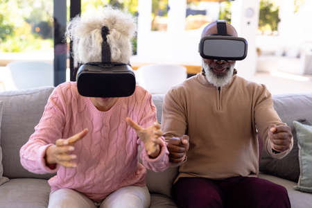 Multiracial excited senior friends enjoying virtual reality simulators on sofa in retirement home. Gesturing, unaltered, creative, futuristic, technology, togetherness, support and assisted living.の写真素材