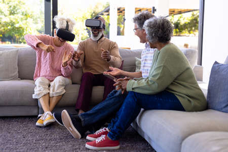Multiracial seniors looking at cheerful friends gesturing while using virtual reality simulators. Retirement home, unaltered, creative, modern technology, togetherness, support, assisted living.の写真素材