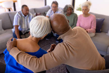 Rear view of multiracial senior man embracing woman in group therapy session at nursing home. Friends, sad, togetherness, comfort, console, unaltered, stress, support, assisted living, retirement.の写真素材