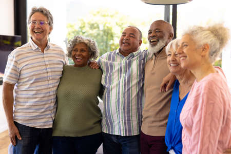 Multiracial senior friends with arms around laughing and looking away while standing in nursing...