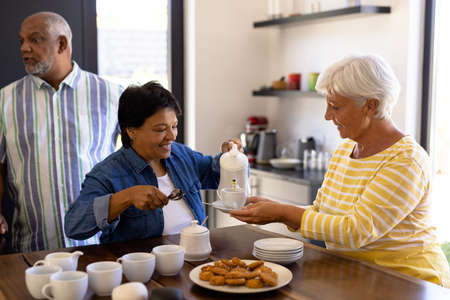 Biracial senior woman serving coffee in cup held by friend at wooden table in nursing home. Snacks,...
