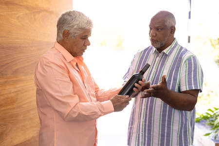 Biracial senior man receiving male friend with champagne bottle while standing in nursing home. Guest, alcohol, entrance, greeting, unaltered, togetherness, support, assisted living, retirement.の写真素材