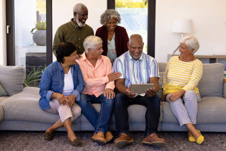 Cheerful multiracial senior friends looking at senior man using digital tablet while sitting on sofa. Nursing home, wireless technology, unaltered, together, support, assisted living, retirement.の写真素材