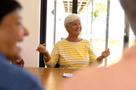 Cheerful biracial senior woman gesturing while playing bingo at dining table in nursing home. Winning, happy, luck, leisure game, unaltered, support, assisted living and retirement concept.の写真素材