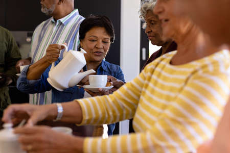 Biracial senior woman serving coffee to multiracial friends while sitting in nursing home. Drink,...